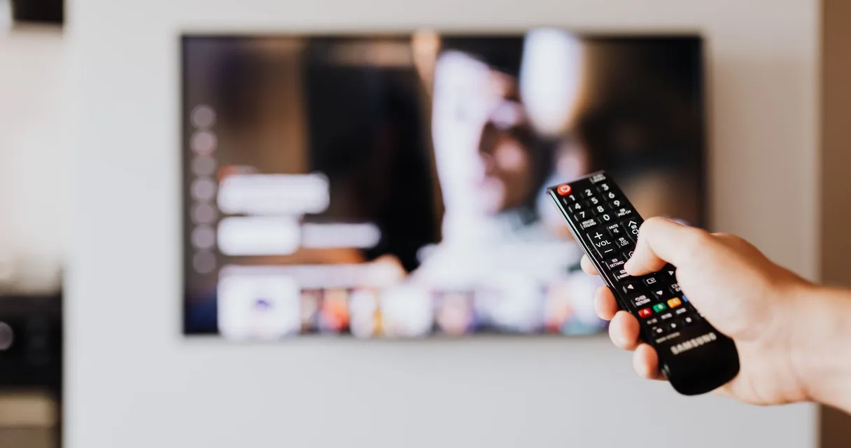 Mão segurando controle remoto diante de uma televisão com menu de programação, ilustrando a lista IPTV