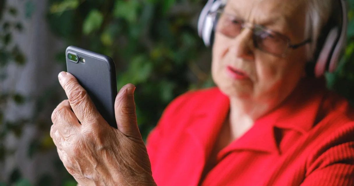Mulher idosa de vermelho usando fones de ouvido e olhando o celular ao ar livre